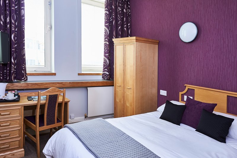 A Kents Hill Park bedroom. A double bed with purple cushions on it is next to a free-standing wooden wardrobe, and a desk with chair. Two large windows have purple curtains.